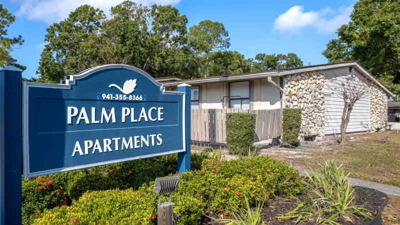 Blue Palm Place Apartments sign with a white swan logo, green landscaping, and a stone-faced building under a blue sky. Photograph.