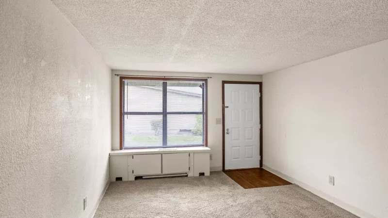 Empty room with white walls, large window, heating unit, and a white door with a mat on light carpet.
