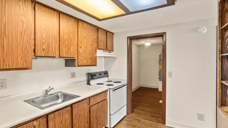 Small kitchen with wooden cabinets, white countertops, stainless steel sink, and white electric stove. Hardwood floor extends to open doorway.