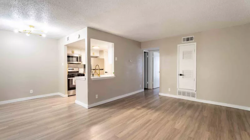 Empty living room with light wood floors, beige walls, and a cutout overlooking a kitchen.