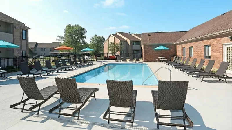 Sunny outdoor swimming pool with numerous lounge chairs, surrounded by apartment buildings under a clear blue sky.