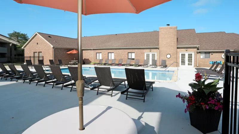 Sunny outdoor swimming pool with lounge chairs, a brick building, and an orange patio umbrella.