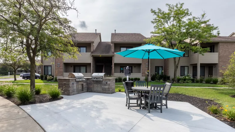 Outdoor patio with a stone grill, a table with chairs, and a turquoise umbrella in front of apartment buildings.