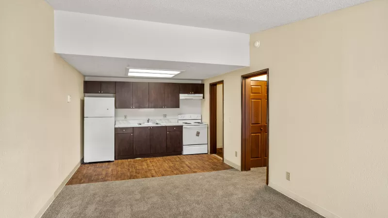 View of a compact apartment kitchen with dark brown cabinets, white appliances, and laminate flooring, opening to a carpeted room.