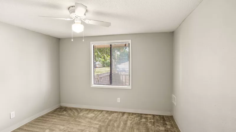 Empty room with light walls, beige carpet, a window with an outdoor view, and a white ceiling fan.