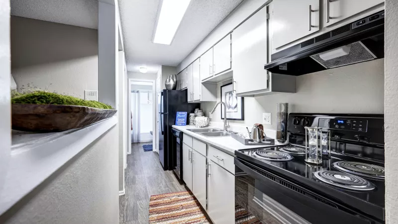 Narrow galley kitchen with white cabinets, black appliances, wood-look floor, and a colorful striped rug.