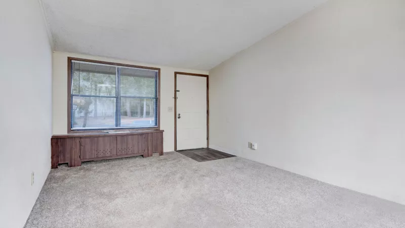 Empty room with beige walls, light carpet, a large window over a wood cabinet, and a white door.