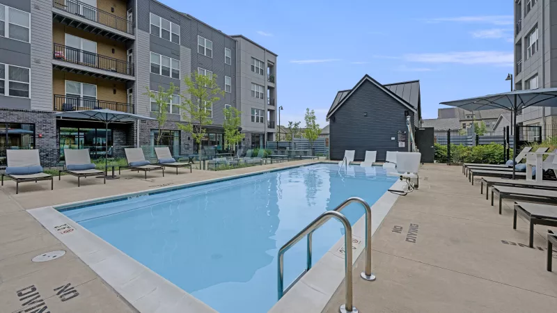 Outdoor swimming pool surrounded by lounge chairs, with modern apartment buildings in the background.