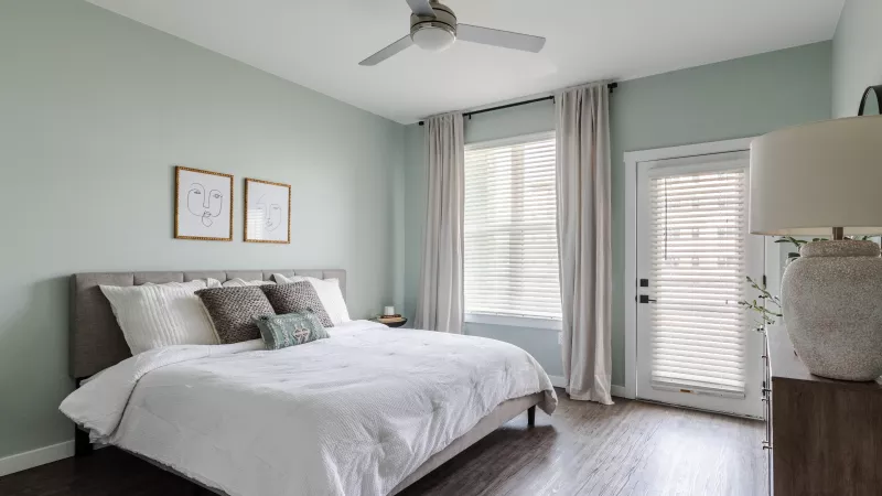 Photograph: Modern bedroom with light green walls, queen bed, ceiling fan, and a glass door to a balcony.