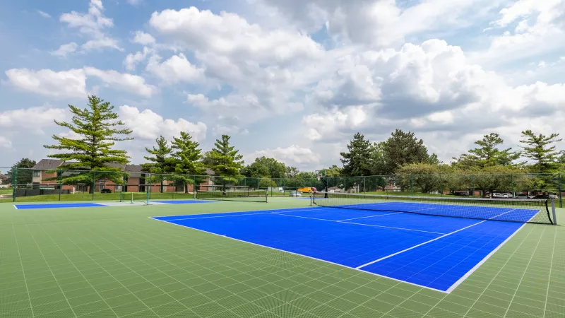 Bright blue tennis court on a green ground, surrounded by trees and buildings, under a blue sky with white clouds.