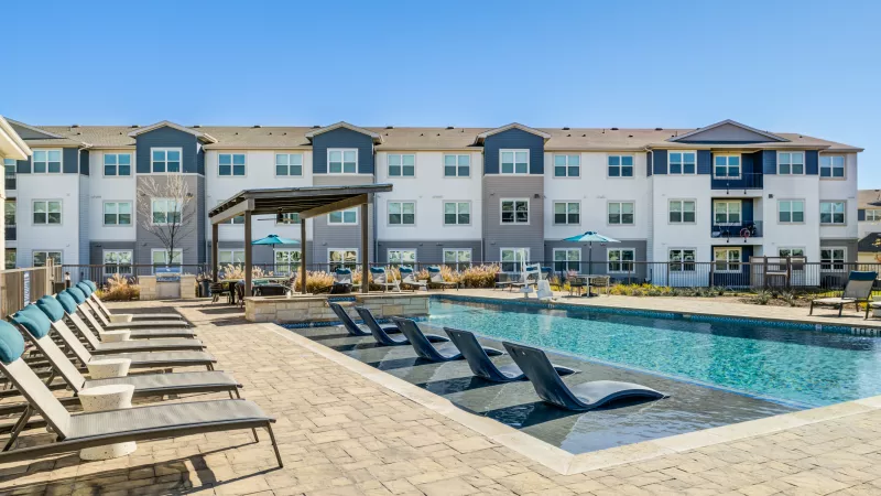 Modern apartment complex with an outdoor swimming pool, lounge chairs, and a clear sunny sky.