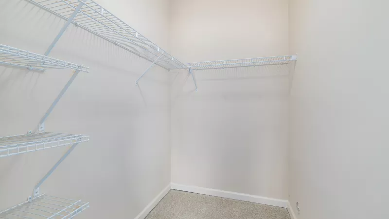 Empty walk-in closet with white wire shelving on three walls, light beige walls, and gray carpet.