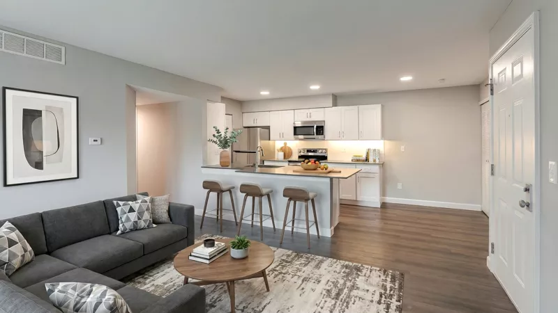 Modern open-plan living room with gray L-sofa, coffee table, and kitchen island with white cabinets and bar stools.