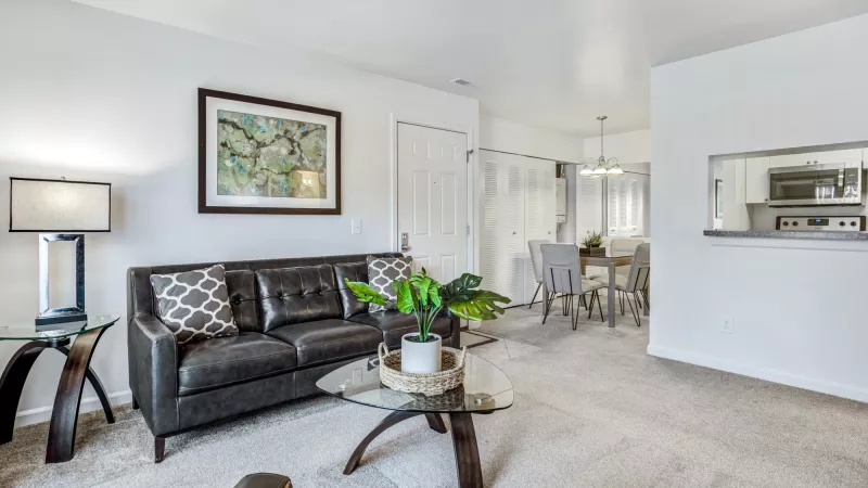 Modern living room with dark leather sofa, glass coffee table, and a lamp. Dining area and kitchen pass-through in background.