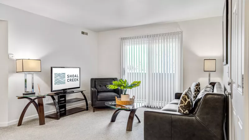 Modern living room with dark gray sofa, black armchair, TV, and glass tables. Sliding door with vertical blinds.