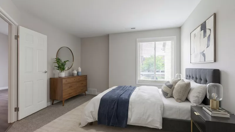 Modern bedroom with a gray upholstered bed, white duvet, blue throw, wooden dresser, round mirror, and a large window.