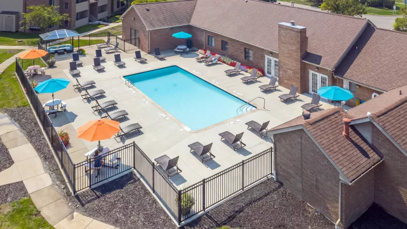 Aerial view of a community pool area with a fence, lounge chairs, blue and orange umbrellas, and a brick building.