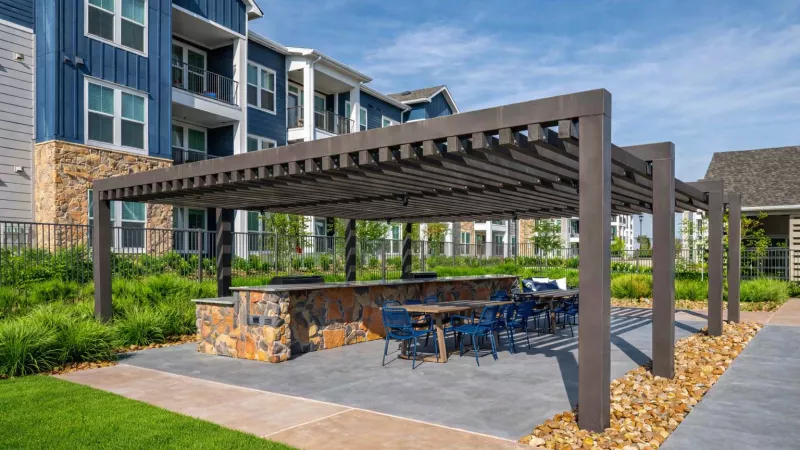 Outdoor patio with a dark pergola shading a stone grill, table, and blue chairs. A modern blue apartment building in the background.