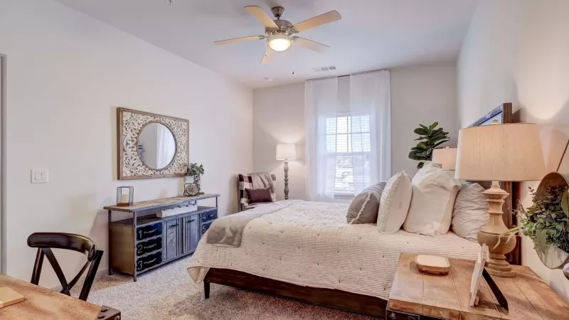 Bright bedroom with a large bed, white bedding, wooden nightstands, a decorative mirror, and a ceiling fan.