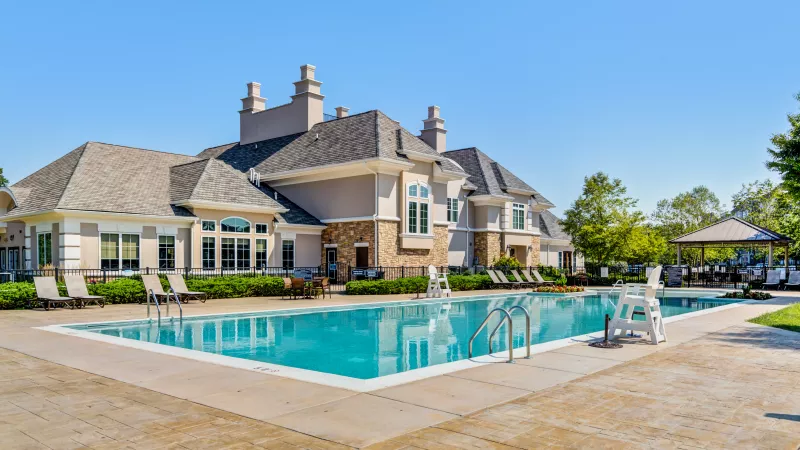Photograph of a luxurious clubhouse with a large swimming pool, lounge chairs, and a gazebo on a sunny day.