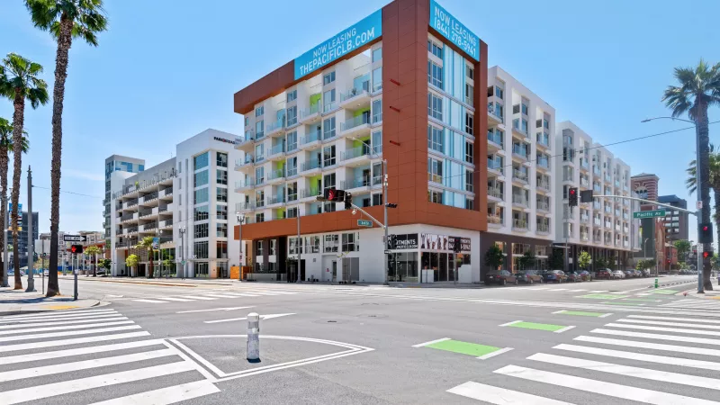 Contemporary brown and white building with balconies at a sunny city intersection with palm trees and a crosswalk.
