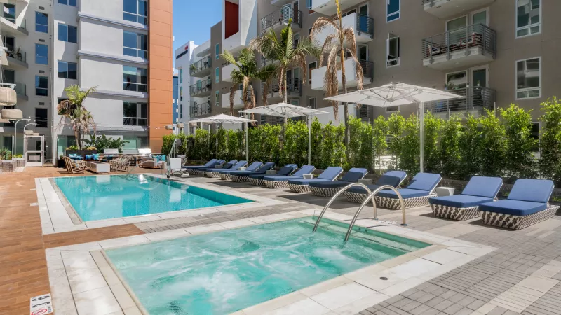 Sunny outdoor pool area with hot tub, blue lounge chairs, palm trees, and modern apartments.