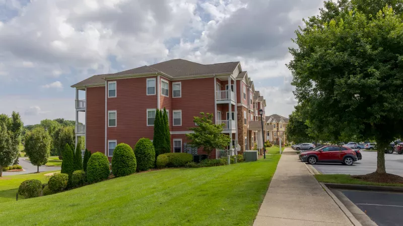 Exterior view of a multi-story red brick apartment building with balconies, green lawn, sidewalk, and parking lot.