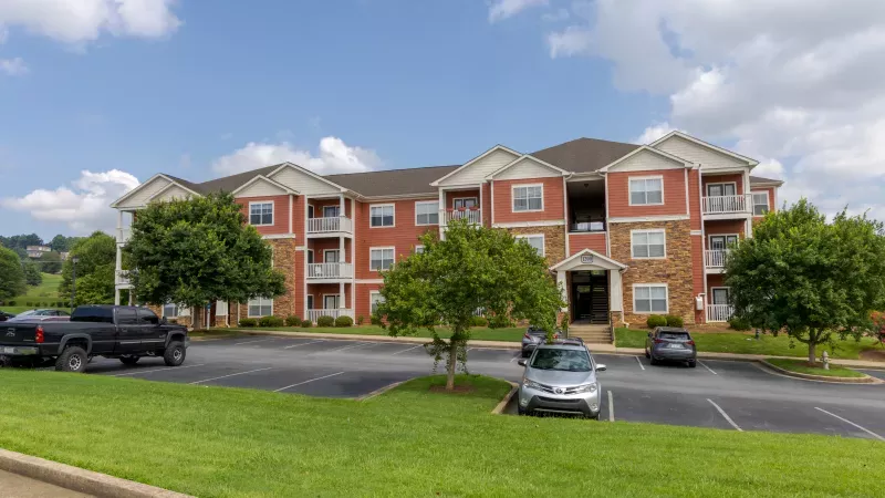 Red-orange apartment building with stone accents, balconies, a parking lot, and green trees under a blue sky.
