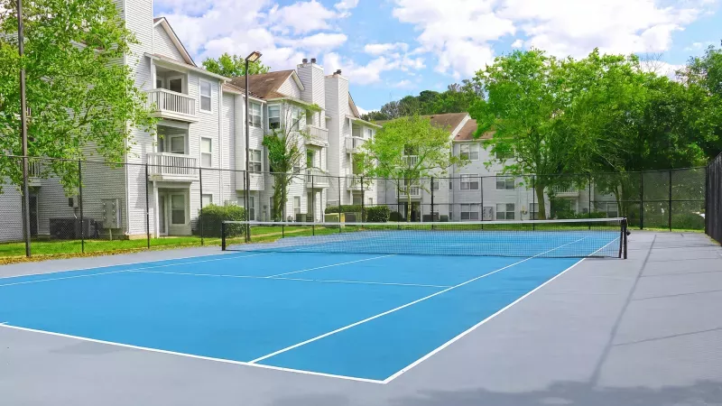 Blue tennis court with white lines and net, surrounded by apartment buildings and green trees under a blue sky.