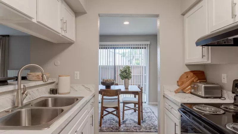 Photograph of a modern white kitchen with marble counters, double sink, and stove, looking into a dining area with a table and glass door.