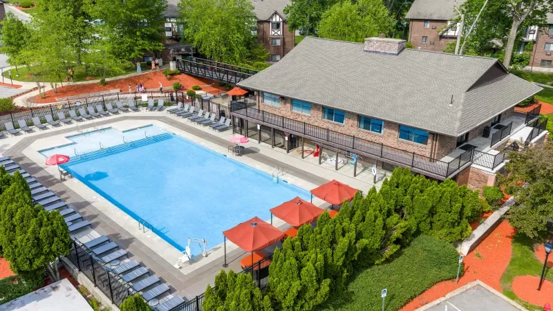 Aerial view of a large outdoor swimming pool with lounge chairs, a clubhouse, red umbrellas, and green landscaping.