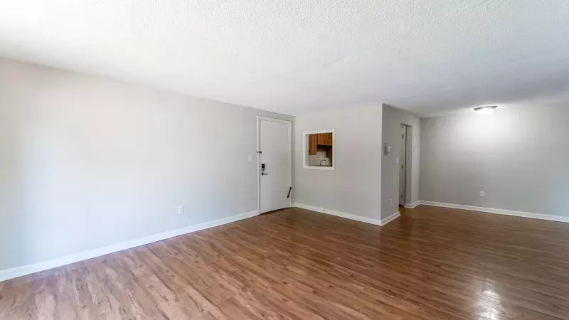 Empty living room with light neutral walls and warm wood floors. A white entry door and small framed art are visible.