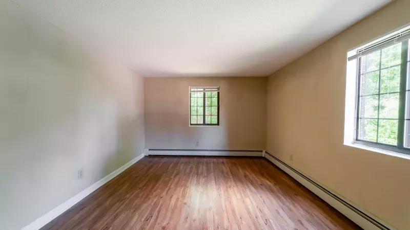 Empty room with warm tan walls, wood-grain floor, and two windows showing green trees.