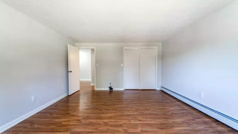 Empty room with light gray walls, wood laminate flooring, an open doorway, closet, and baseboard heater.