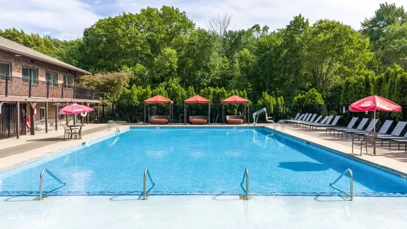 Sunny outdoor pool with blue water, lounge chairs, red umbrellas, and cabanas. A building and trees are in the background.