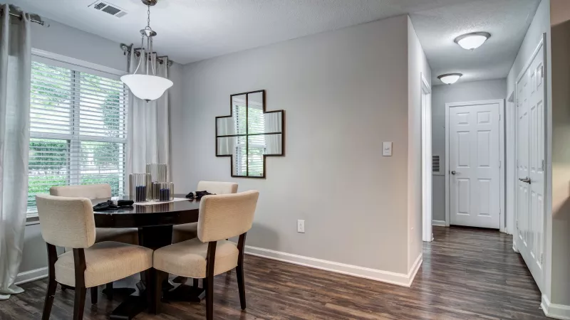 Modern dining room features a dark round table, four beige chairs, and a geometric wall mirror. Bright window and hallway.