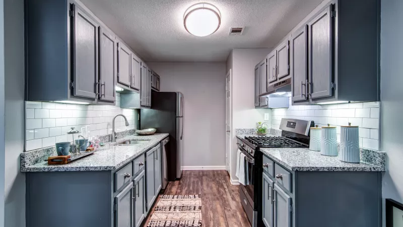 Modern galley kitchen with light blue-gray cabinets, white subway tile, speckled counters, and dark wood-look flooring.