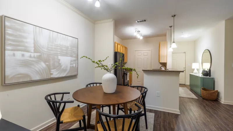 Modern dining room with a round wooden table, four black chairs, and a white vase with green branches. Kitchen in background.