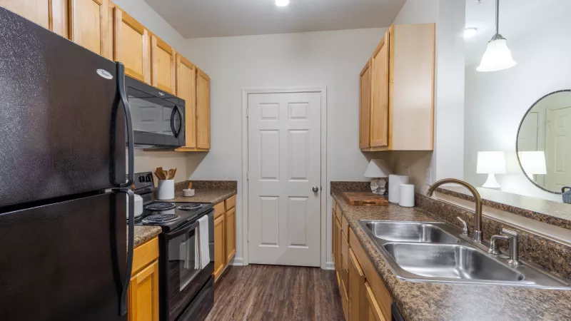 Modern galley kitchen with black appliances, light wood cabinets, dark speckled counters, and a stainless steel double sink.