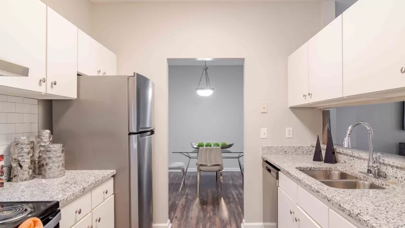 Modern kitchen with white cabinets, granite counters, stainless steel fridge, leading to a dining room.