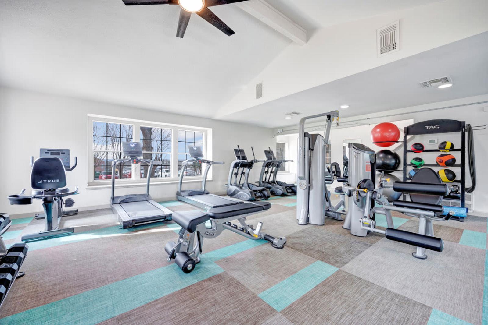 Bright and spacious fitness center equipped with modern exercise machines, including treadmills, ellipticals, and strength-training equipment. The space features large windows, colorful flooring, and ceiling fans, creating an inviting and energizing workout environment.