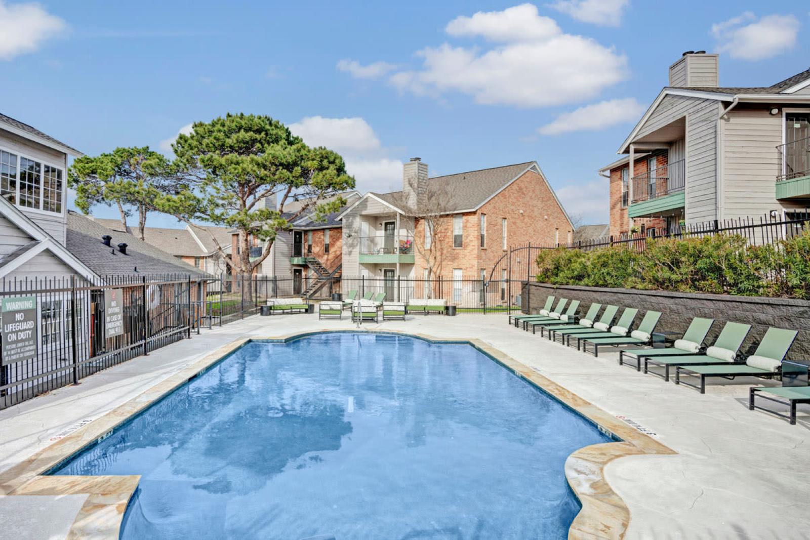  Refreshing outdoor pool with lounge chairs surrounding the clear blue water, set against a backdrop of modern apartment buildings and green trees. The bright blue sky enhances the relaxing atmosphere of the pool area.