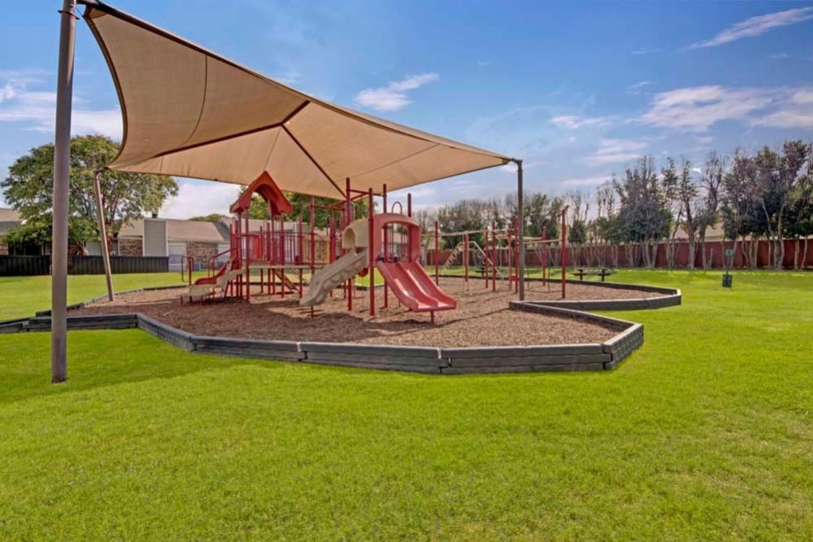 A vibrant playground with red slides and climbing equipment shaded by a large canopy in a green park setting.