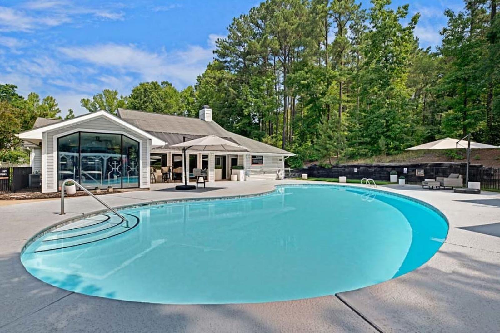 An outdoor swimming pool with forested surroundings and a clubhouse in the background.
