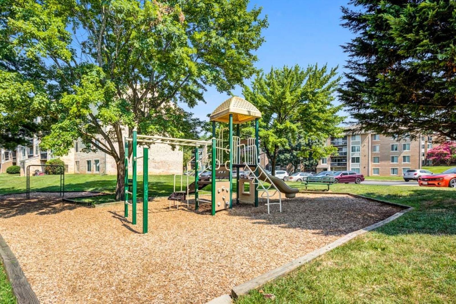 A vibrant playground with slides and climbing structures surrounded by mature trees and apartment buildings in the background.