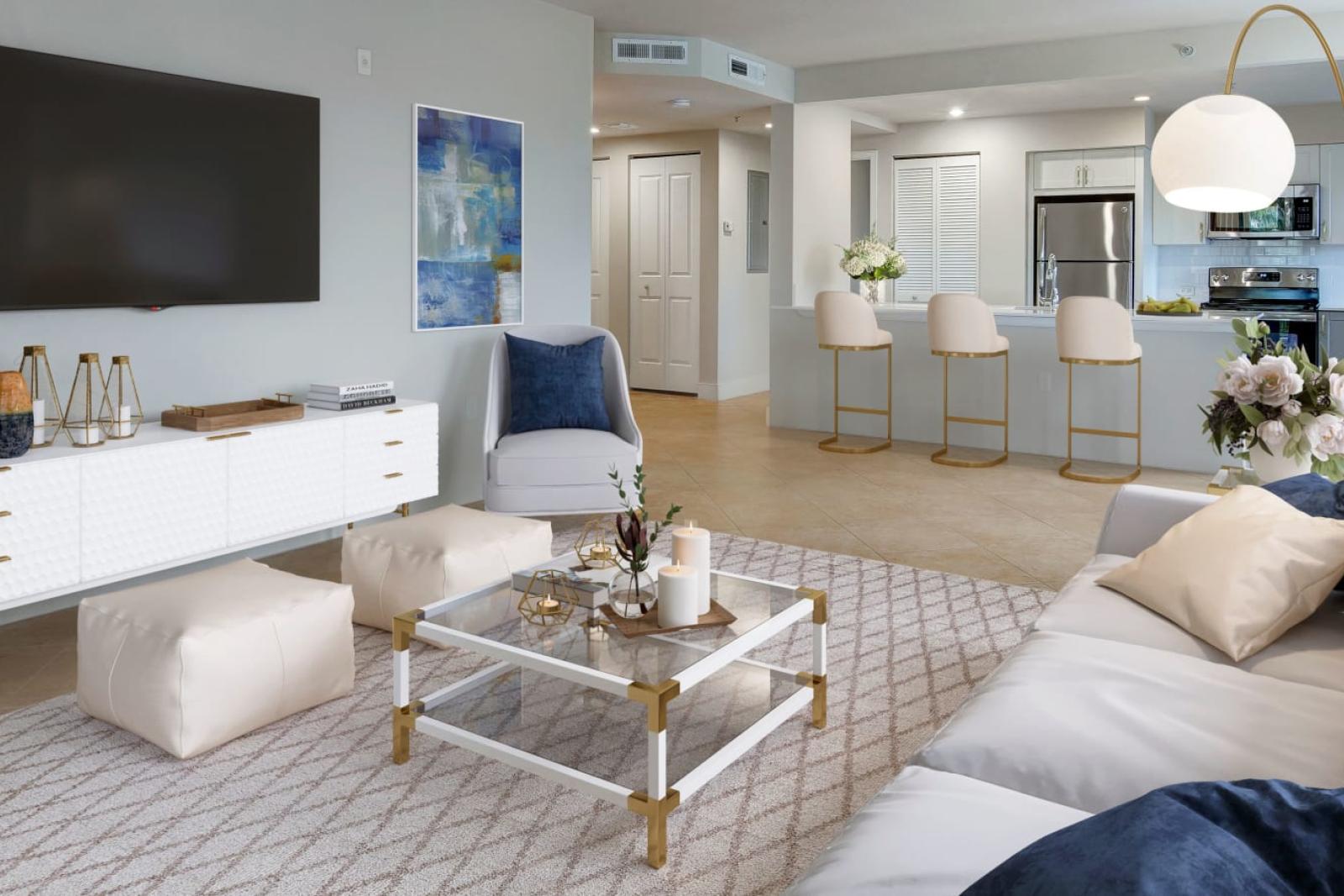 A bright and modern living room with white and gold decor, a mounted TV, and a view into an open-concept kitchen with a bar area.