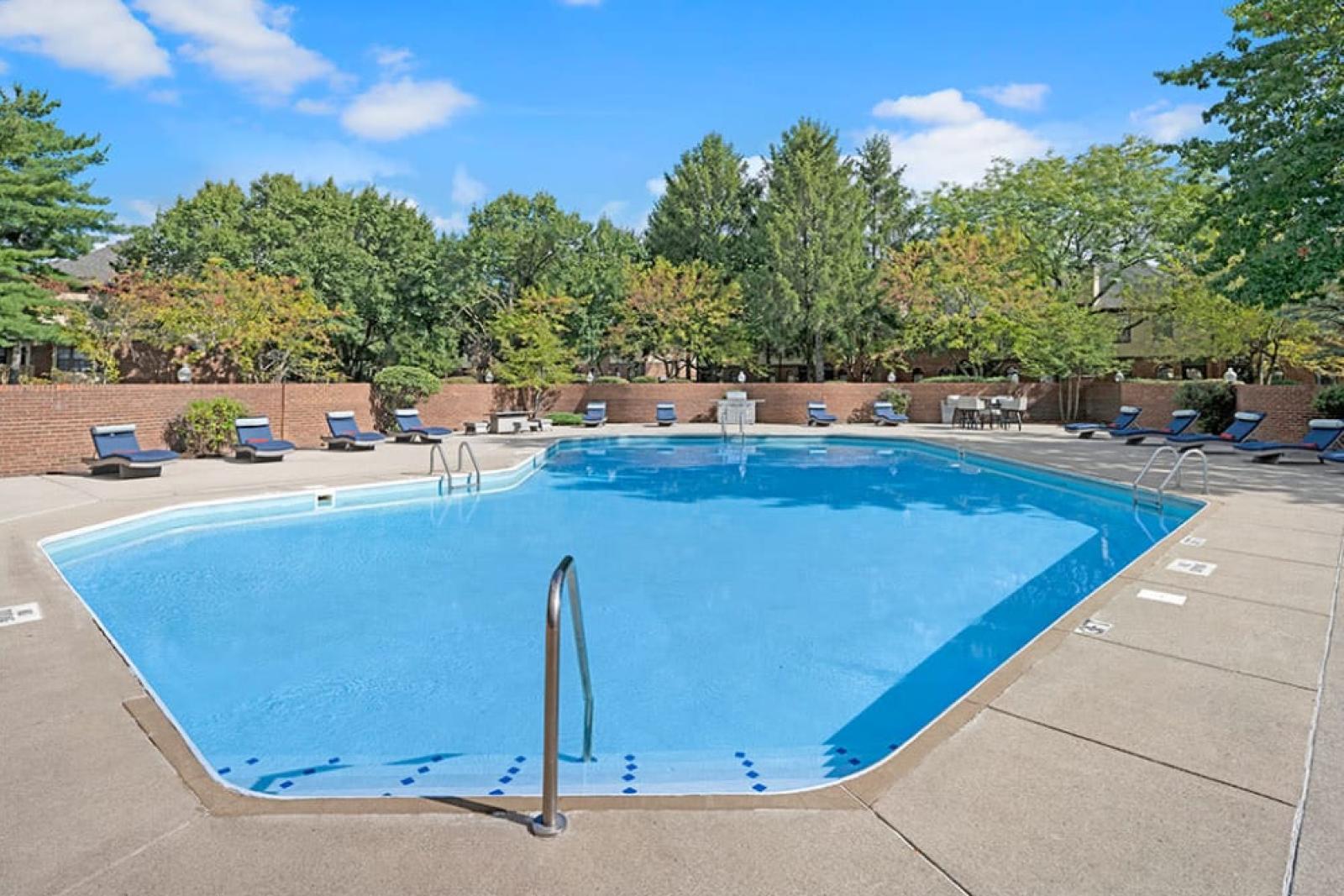 A large outdoor swimming pool surrounded by lounge chairs and mature trees under a clear blue sky.