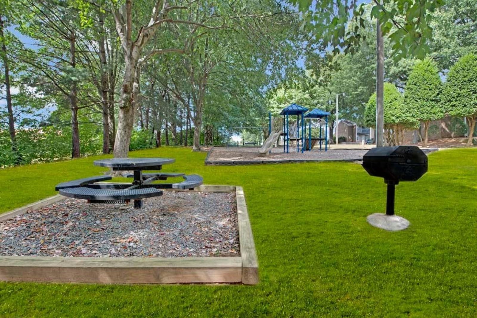 A picnic area with a circular table and a grill adjacent to a playground in a wooded setting.