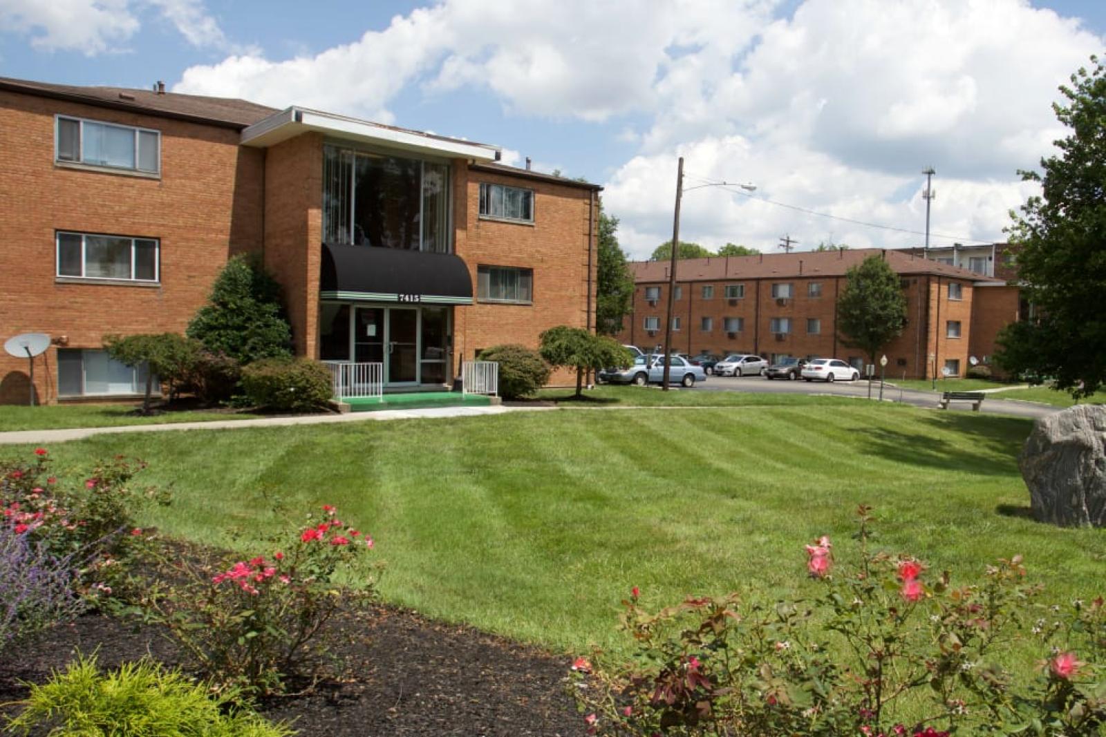 An exterior view of a brick apartment complex with well-maintained green lawns and colorful flowerbeds.