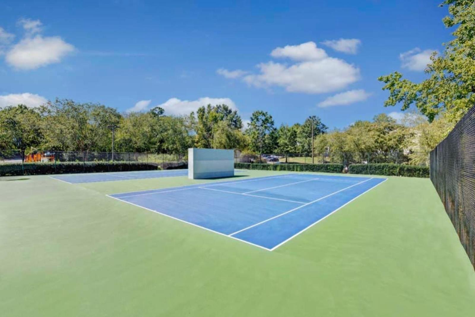 A vibrant blue tennis court sits under a clear blue sky, surrounded by lush greenery.